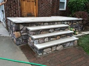 Stone steps and a porch with a smooth concrete top lead to a wooden front door of a brick house. A small garden and a green hose are visible beside the steps.