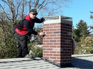 A person wearing black clothes and a red vest works on a brick chimney on a rooftop, holding a tool and inspecting or repairing the chimney cap. Trees and a clear blue sky are in the background.