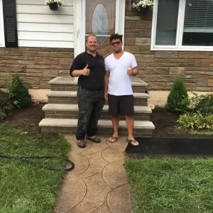 Two men standing and smiling on a sidewalk in front of a house, both giving thumbs up. One wears black work clothes and the other wears a white t-shirt, shorts, and flip-flops. Manicured bushes and stone steps are behind them.