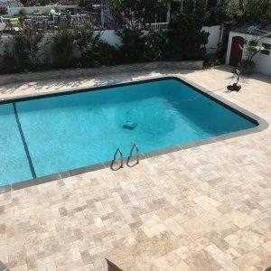 A rectangular swimming pool with clear blue water is surrounded by light-colored stone tiles expertly installed by a masonry contractor Long Island, NY. A pool cleaning robot is submerged, another on the deck, with lush plants and a white fence in the background.