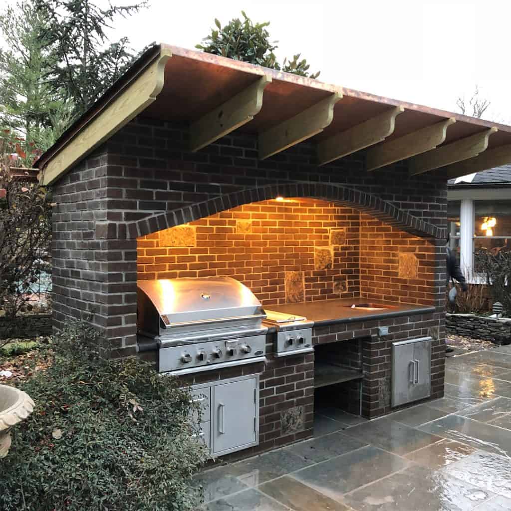 An outdoor kitchen area on a gray stone patio. It features a large, built-in, dark brick structure with a sloped wooden roof. A large stainless steel grill and a countertop with an integrated sink are built into the brick.