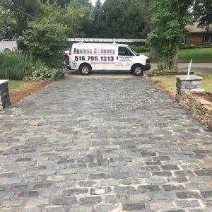 A paved stone driveway leads to a parked white van with "Ageless Chimney" and a phone number on the side. The driveway is bordered by stone pillars and greenery on both sides.
