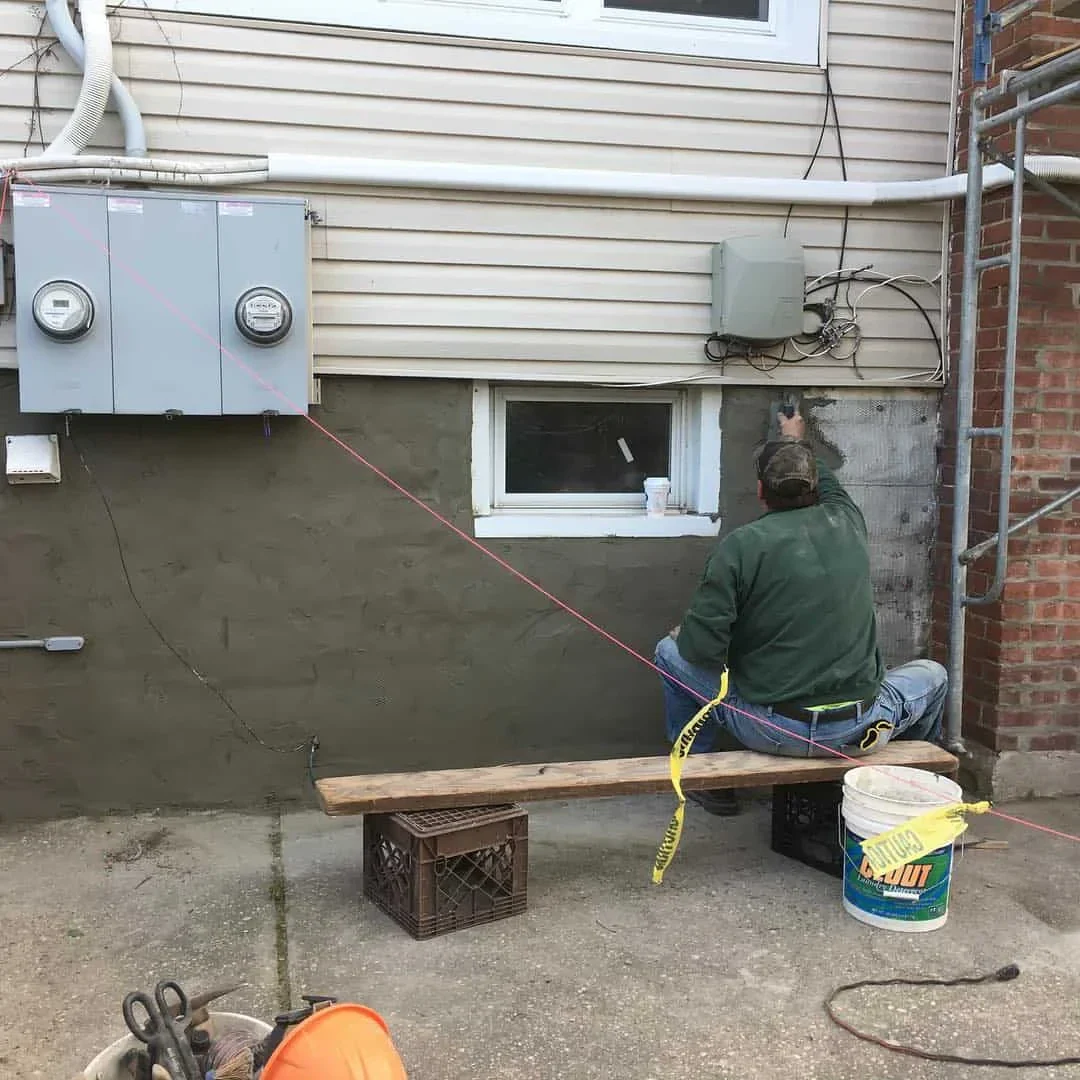 A person in NY sits on a wooden plank supported by crates, working as a masonry contractor Long Island on an exterior wall, surrounded by utility boxes, wires, construction materials, and caution tape.