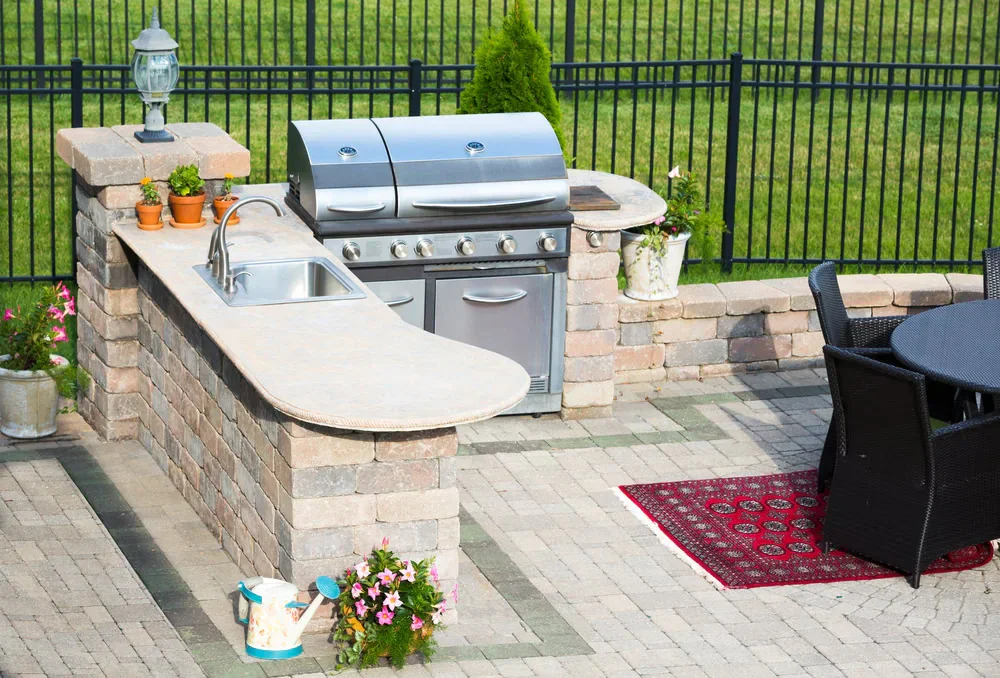 Outdoor kitchen with a stone counter crafted by a top masonry contractor Long Island, built-in sink, stainless steel grill, potted plants, and a round dining table on the patio, overlooking green grass and a black metal fence in NY.